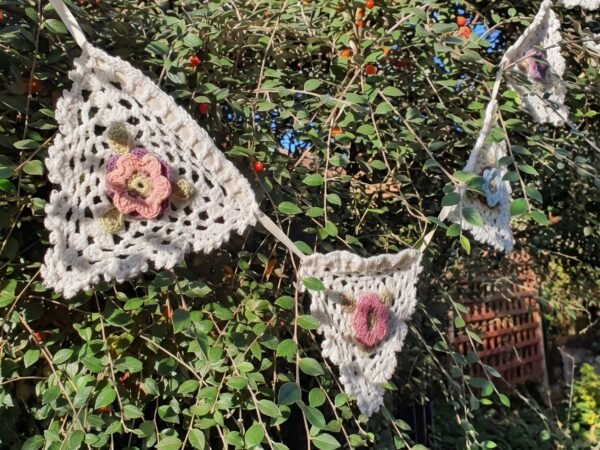 Handcrafted cream crochet bunting featuring triangular openwork flags, each decorated with a 3D floral accent in pink, purple, and blue, hanging against lush green garden foliage.