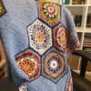 Handmade crochet blanket featuring colorful floral hexagon motifs surrounded by soft blue textured stitching, draped over an armchair.