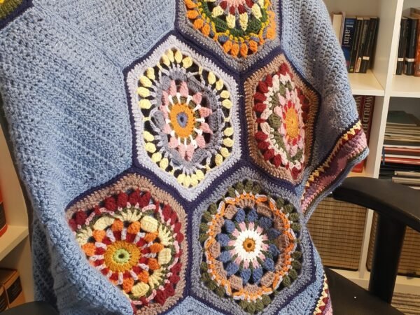 Handmade crochet blanket featuring colorful floral hexagon motifs surrounded by soft blue textured stitching, draped over an armchair.