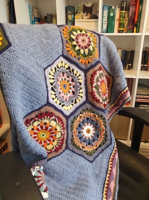 Handmade crochet blanket featuring colorful floral hexagon motifs surrounded by soft blue textured stitching, draped over an armchair.